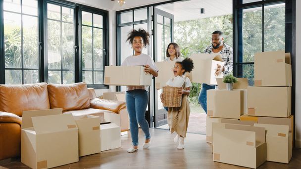 Happy African American young family bought new house. Mom, Dad, and child smiling happy hold cardboard boxes for move object walking into big modern home. New real estate dwelling, loan and mortgage. - Фото, зображення