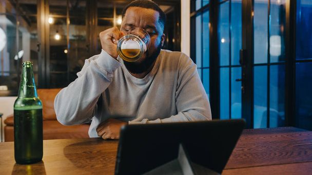 Középkorú afro-amerikai férfi sörivás szórakozás boldog éjszakai party esemény online ünneplés videohívás a nappaliban otthon. Társadalmi távolság, karantén a koronavírus megelőzésére. - Fotó, kép