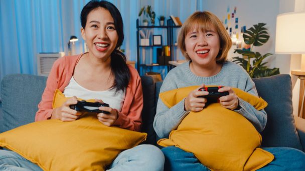 Лесбійські ЛГБТ-жінки грають вдома у відеоігри. Молода азіатка, що користується бездротовим контролером, веселиться вночі на дивані у вітальні. Вони чудово проводять час, святкують свято.. - Фото, зображення