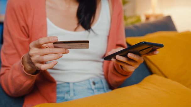 Nacht schöne lächelnde asiatische Dame verwenden Handy bestellen Online-Shopping-Produkt mit Kreditkarte auf dem Sofa im Wohnzimmer. Bleiben Sie zu Hause, Selbst Quarantäne Aktivität, Spaß Aktivität für covid Quarantäne. - Foto, Bild