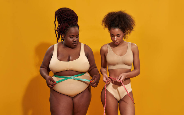 Two young african american women with different body size measuring their waist using metric tape measure after a diet, posing together isolated over orange background - Foto, imagen