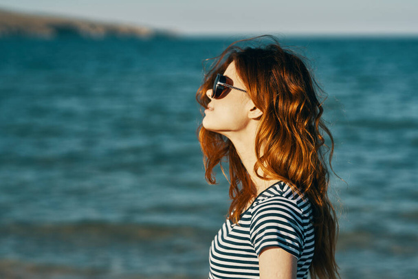 mujer pelirroja en gafas de sol en la playa cerca del mar azul y las montañas en el fondo - Foto, imagen