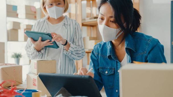 Genç Asyalı iş kadınları hisse senetlerinde yüz maskesi ürün satın alma siparişi takıyor ve merkez ofisindeki tablet bilgisayarlara kaydediliyor. Küçük işletme sahibi, çevrimiçi pazar teslimatı, yaşam tarzı serbest konsepti - Fotoğraf, Görsel