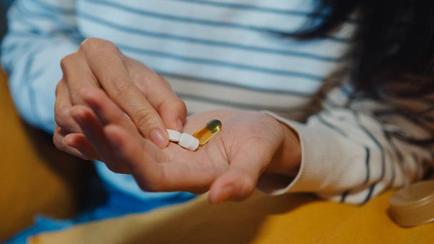 Zieke Aziatische jongedame met pil neemt medicijnen op de bank in de woonkamer thuis 's nachts. Meisje dat drugs gebruikt na doktersvoorschrift, quarantaine thuis, Coronavirus sociaal afstandelijk gezondheidsconcept. - Foto, afbeelding