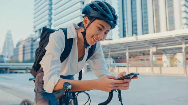 バックパック付きの笑顔アジアのビジネスマンの女性は、自転車がオフィスで働くために行くと街の屋台でスマートフォンルックカメラを使用します。スポーツ少女は仕事に電話を使う。通勤、通勤、通学. - 写真・画像