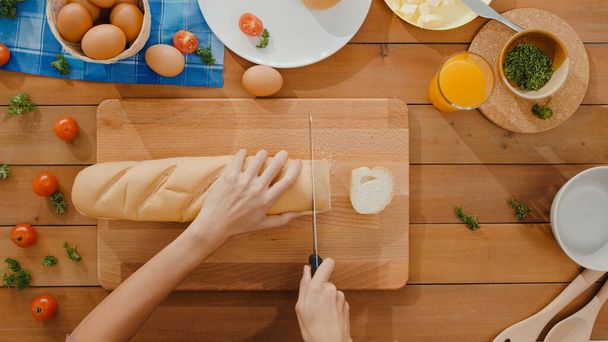 Τα χέρια της νεαρής Ασιάτισσας σεφ κρατούν μαχαίρι κομμένο ψωμί ολικής αλέσεως σε ξύλινη σανίδα στο τραπέζι της κουζίνας στο σπίτι. Φρέσκο σπιτικό ψωμί, υγιεινό φαγητό και παραδοσιακή έννοια αρτοποιίας. Φωτογραφία με πάνω όψη. - Φωτογραφία, εικόνα