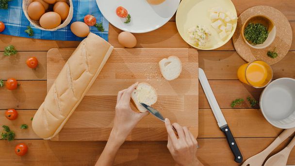 Młoda azjatycka kucharka rozkładała masło na chlebie żytnim z metalowym nożem na drewnianej desce na stole kuchennym w domu. Świeże domowe pieczywo, zdrowe jedzenie i tradycyjna piekarnia. Widok z góry. - Zdjęcie, obraz