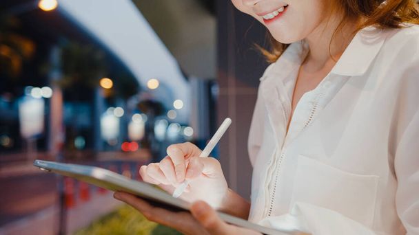 Moda bürosu kıyafetleri içinde başarılı genç Asyalı iş kadını dijital tablete yazmak için akıllı kalem teknolojisi kullanırken gece şehir çağında açık havada tek başına oturuyor. İş konsepti.. - Fotoğraf, Görsel