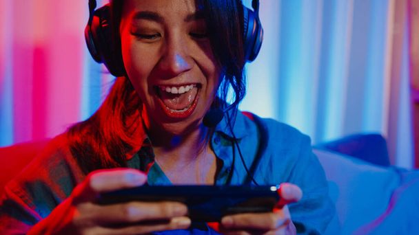 Happy Asia tyttö gamer kulumista kuulokkeet kilpailu videopeli verkossa älypuhelin innoissaan puhua ystävän kanssa istua sohvalla värikäs neon valot olohuone kotona, Home karanteeni toiminta käsite. - Valokuva, kuva