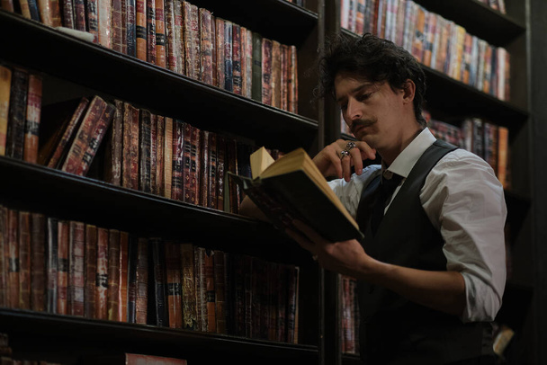 задумчивый человек стоит ночью в темной библиотеке и держит винтажную книгу. стильный мужчина с усами выпрямляет браслет. - Фото, изображение