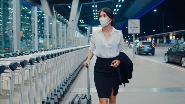 Asyalı iş kızı varış yerine giderken maske takıyor ve iç havaalanındaki garaj kapısının önünde kalkıyor. İş seyahati covid salgını, iş seyahati sosyal uzaklık kavramı. - Fotoğraf, Görsel