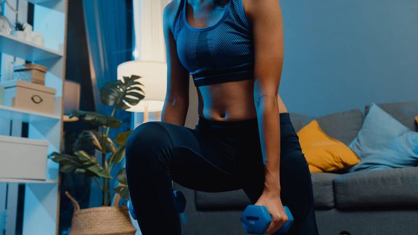 Young Asia lady in sportswear doing squat with dumbbells in outstretched arms exercise working out in living room at home at night. Sport and recreation activity, quarantine for corona virus concept. - Fotografie, Obrázek