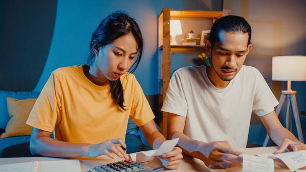 ストレスアジアのカップルの男性と女性は、家庭での金融経済危機時の家計、債務、費用を計算するために電卓を使用します夜。結婚資金のトラブル、家族の予算計画の概念. - 写真・画像
