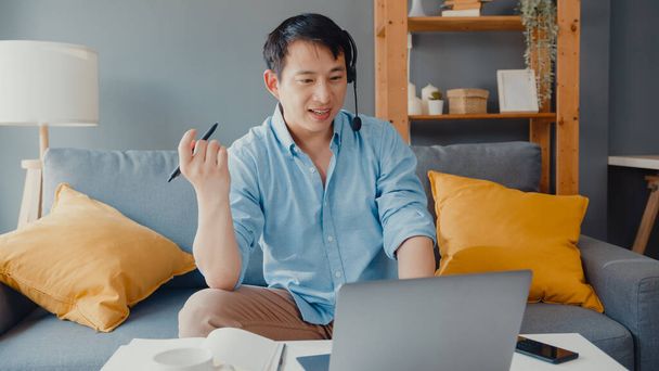Молодой азиатский бизнесмен носит наушники, используя ноутбуки, чтобы поговорить с коллегами о плане в видеозвонке во время работы из дома в гостиной. Самоизоляция, социальное дистанцирование, карантин для профилактики ковидов. - Фото, изображение