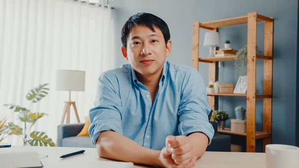 自宅のリビングルームで働いている間、若いアジアのビジネスマンは、ビデオ通話会議の計画について同僚にコンピュータのラップトップトークを使用しています。自己分離、社会的距離、コロナウイルスの隔離. - 写真・画像