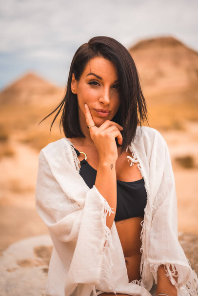 Sensual look of a young brunette Caucasian girl in a white dress sitting in a beautiful desert - Photo, Image
