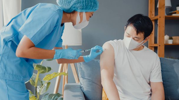 Jovem enfermeira da Ásia dando vacina antivírus Covid-19 ou gripe disparada para o paciente masculino usar proteção máscara facial contra a doença viral sentar no sofá na sala de estar em casa. Conceito de vacinação. - Foto, Imagem