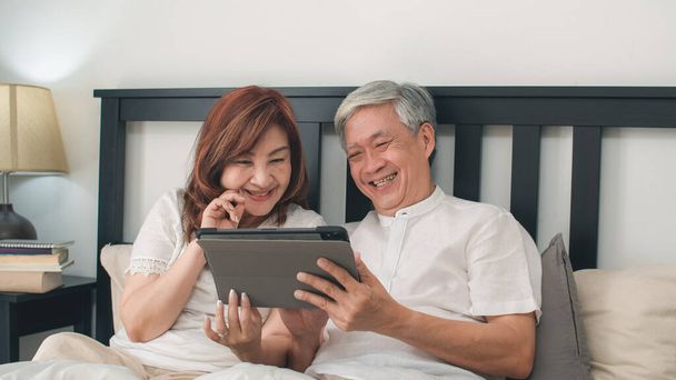 Asiatisches Seniorenpaar mit Tablet zu Hause. Asiatische Senioren chinesische Großeltern, Mann und Frau glücklich nach dem Aufwachen, Film auf dem Bett liegend im Schlafzimmer zu Hause in der Morgen-Konzept. - Foto, Bild