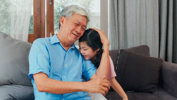 Dziadek z Azji rozmawia z wnuczką w domu. Senior chiński, dziadek szczęśliwy relaks z młodą wnuczką dziewczyna za pomocą czasu rodzinnego relaks z młodą dziewczyną dziecko leżące na kanapie w salonie. - Zdjęcie, obraz