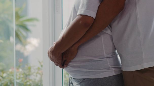 Junge asiatische Schwangere umarmen und halten Bauch im Gespräch mit ihrem Kind. Mama und Papa glücklich lächelnd friedlich, während Pflege Baby, Schwangerschaft in der Nähe Fenster im Wohnzimmer zu Hause Konzept. - Foto, Bild