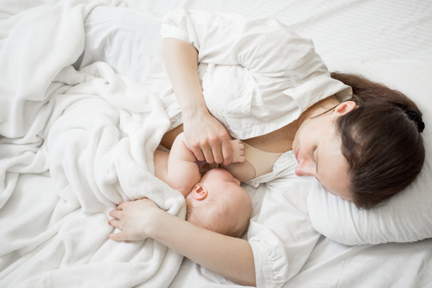 Breastfeeding in a comfortable lying position. Mom with a newborn lie at home in bed on white linens. A lifestyle moment. Family time. Caucasian mother with newborn baby. - Photo, Image