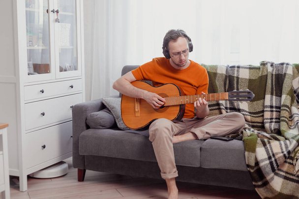 Nuori valkoihoinen mies kuulokkeissa soittaa kitaraa kotona sohvalla. Harjoittele musiikkia, elämäntapa valokuva - Valokuva, kuva