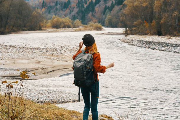 путешественница в шляпе в свитере с рюкзаком жестикулирует руками и расслабляется на берегу реки в горах - Фото, изображение