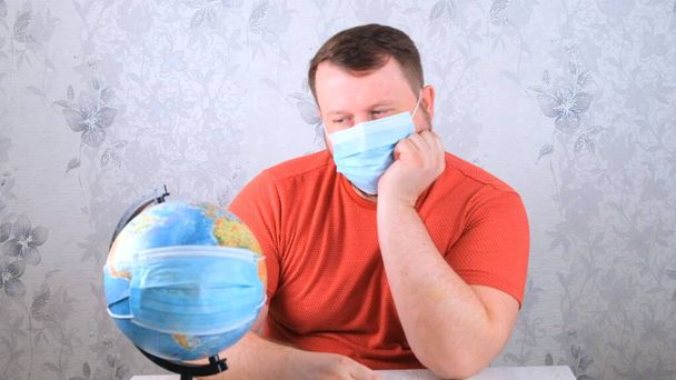 man is sad about the illnesses on the plan and the lack of travel. male looks globe in protective mask and pandemic vaccine. Corona virus treatment concept, injection, clinical trials during a - Zdjęcie, obraz
