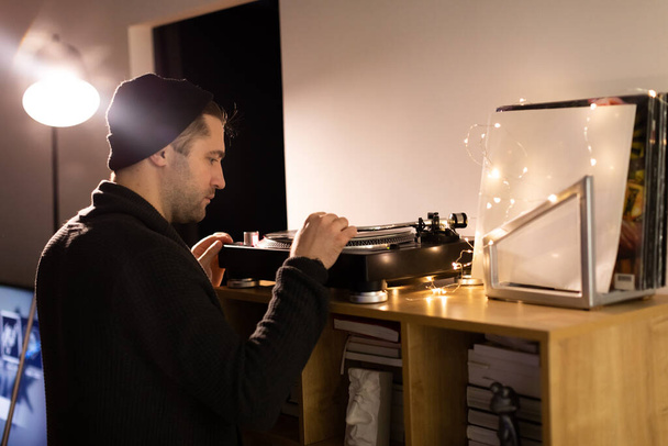 Young unshaven handsome hipster guy puts tonearm on vinyl record on turntable - Photo, Image