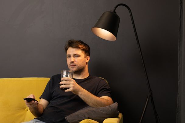 Young unshaven guy watching movie on TV on sofa with drink and remote control - Valokuva, kuva