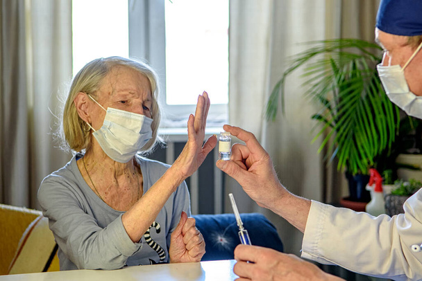 Ένας φροντιστής γιατρός βοηθά μια αρνητική ηλικιωμένη γιαγιά 85 ετών - ένας ασθενής στο σπίτι ή στο νοσοκομείο. Η έννοια της υγειονομικής περίθαλψης για τους ηλικιωμένους. Άρνηση εμβολιασμού - Φωτογραφία, εικόνα