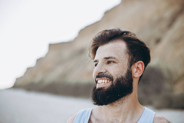 portrait of a stylish young man with a beard and a face on which the sea sand on the background of the sea shore - Foto, Bild