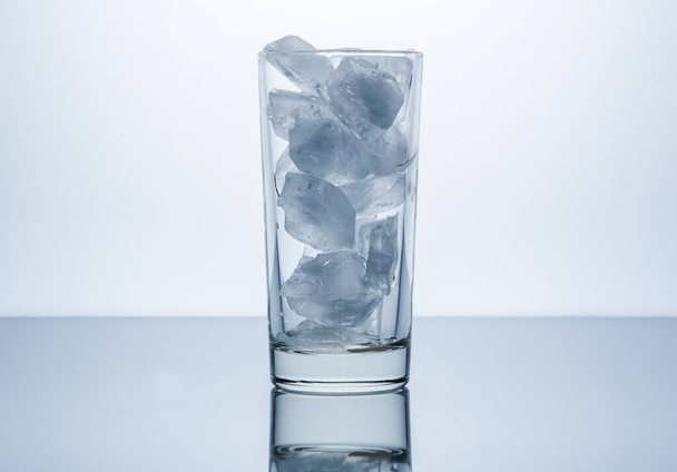 Un vaso transparente lleno de cubitos de hielo se ilumina sobre un fondo blanco. - Foto, Imagen