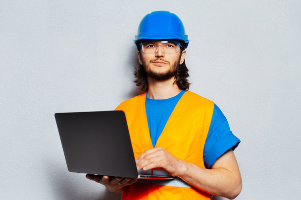 Portret van een jonge zelfverzekerde man, bouwvakker die veiligheidsuitrusting draagt, met laptop op de achtergrond van een grijze muur. - Foto, afbeelding