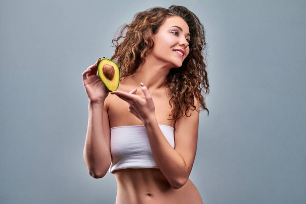 Portrait of a beautiful young woman smiling and holding half an avocado over the side of her face isolated on grey - Photo, image