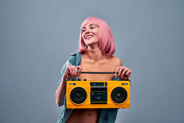 Young sexy smiling woman with pink hair in denim jacket on a naked body holding big old boom box. Isolated grey background - Foto, Imagem