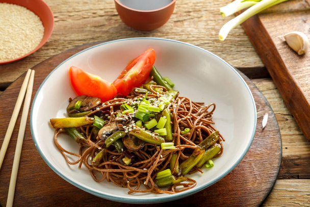 Soba with mushrooms, green beans and sesame seeds in a plate on a stand on a wooden table next to chopsticks and soy sauce and garlic on stands. - Photo, image