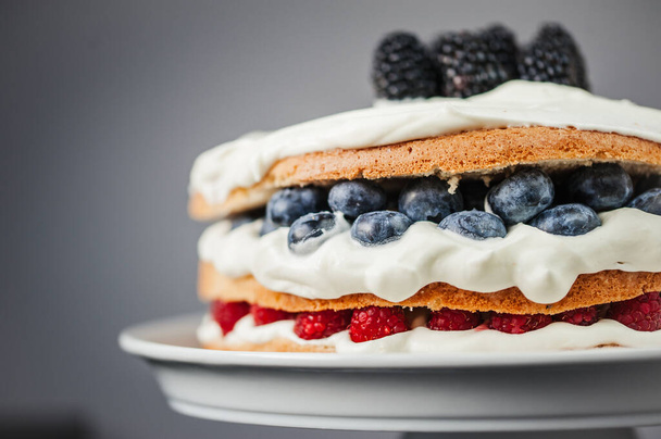 Half of layered sponge cake garnished with white cream frosting and blueberries, raspberries, blackberries, close up on grey background - Foto, afbeelding