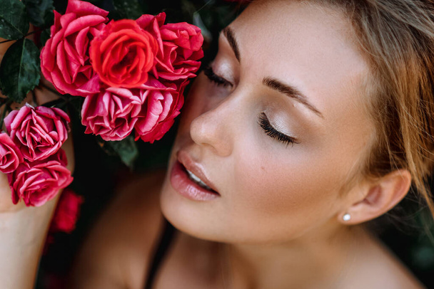 Outdoor photo of romantic young woman in rose garden. girl with red lips in a black dress . Young model on a background of a bush of roses. Stylish woman. - 写真・画像