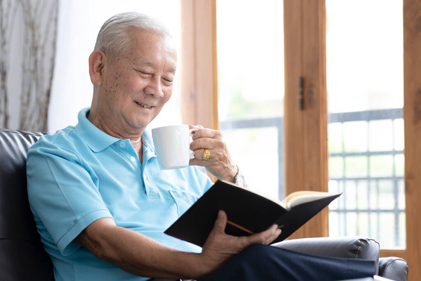 ソファに座ってリビングで面白い本を読んでいる高齢者の男性をリラックス。退職生活の概念. - 写真・画像