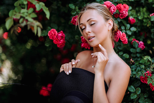 Εξωτερική φωτογραφία της ρομαντικής νεαρής γυναίκας στον κήπο με τα τριαντάφυλλα. κορίτσι με κόκκινα χείλη σε μαύρο φόρεμα. Νεαρό μοντέλο με καπέλο σε φόντο τριανταφυλλιάς. Κομψή γυναίκα.. - Φωτογραφία, εικόνα
