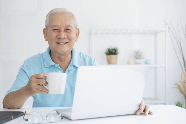 Щасливий старший чоловік працює на ноутбуці і тримає чашку кави вдома
 - Фото, зображення