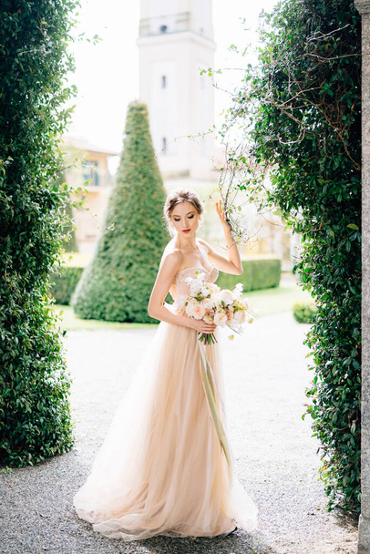 Νύφη σε ένα φόρεμα με ένα μπουκέτο λουλούδια χαμήλωσε το κεφάλι της με φόντο το πράσινο κισσό και αρχαία κτίρια. Λίμνη Κόμο, Ιταλία - Φωτογραφία, εικόνα