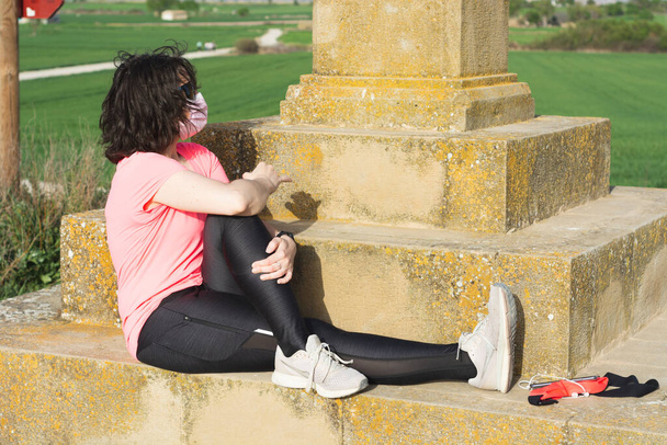 Πορτρέτο της αθλήτριας γυναίκα στο πεδίο κάνει πόδι τέντωμα με ροζ υγιεινή μάσκα. - Φωτογραφία, εικόνα