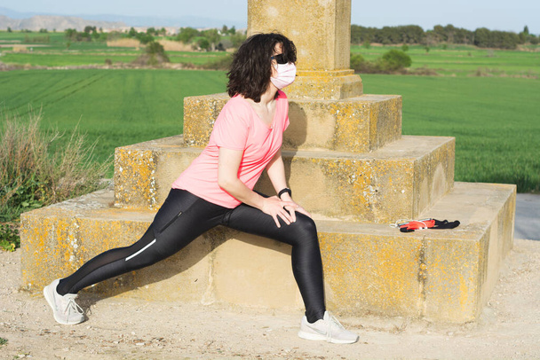 Portrait of athlete woman in the field doing leg stretching with pink hygienic mask. - Photo, Image