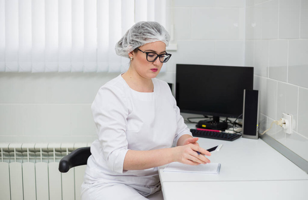 doctora con gafas y un traje médico blanco se sienta en un escritorio con un teléfono y un cuaderno en el consultorio de la clínica - Foto, Imagen