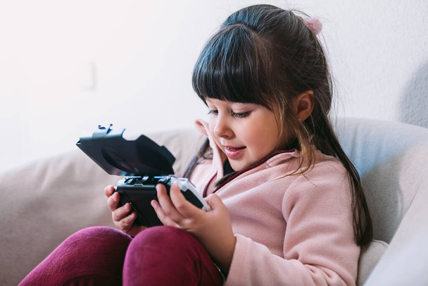 Little black haired girl wearing a pink sweater playing a video game with a portable video console on a sofa - Фото, зображення