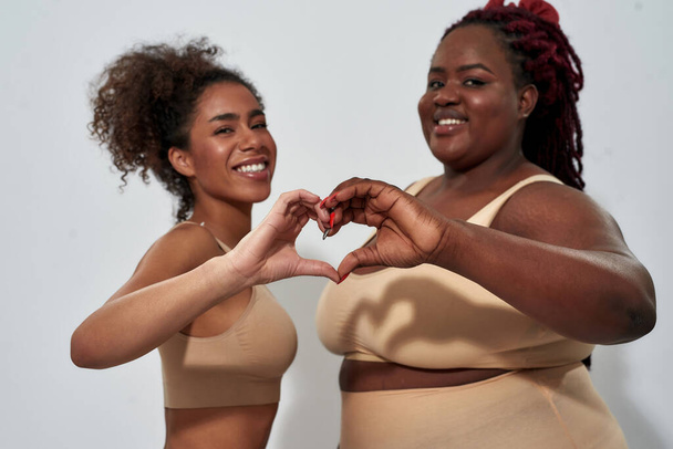 Slim and overweight african american women in underwear with different body size smiling at camera, showing heart love sign, posing isolated over gray background - Foto, Bild
