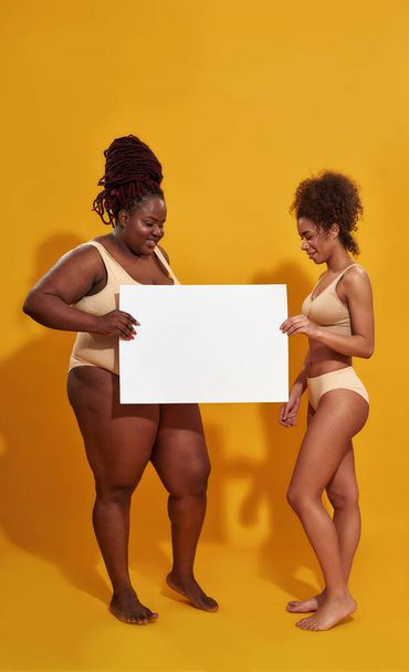 Foto completa de dos alegres mujeres afroamericanas en ropa interior con diferente peso corporal sonriendo, sosteniendo banner en blanco para su texto, de pie aislado sobre fondo amarillo - Foto, imagen