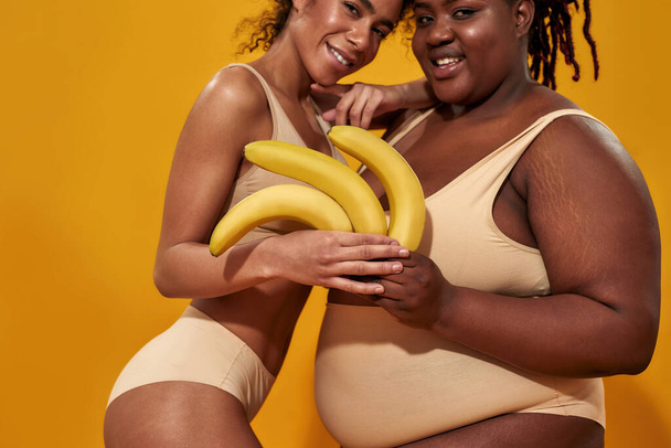 Cropped shot of smiling young african american women in underwear with different body size holding ripe yellow bananas, posing together isolated over orange background - Photo, Image